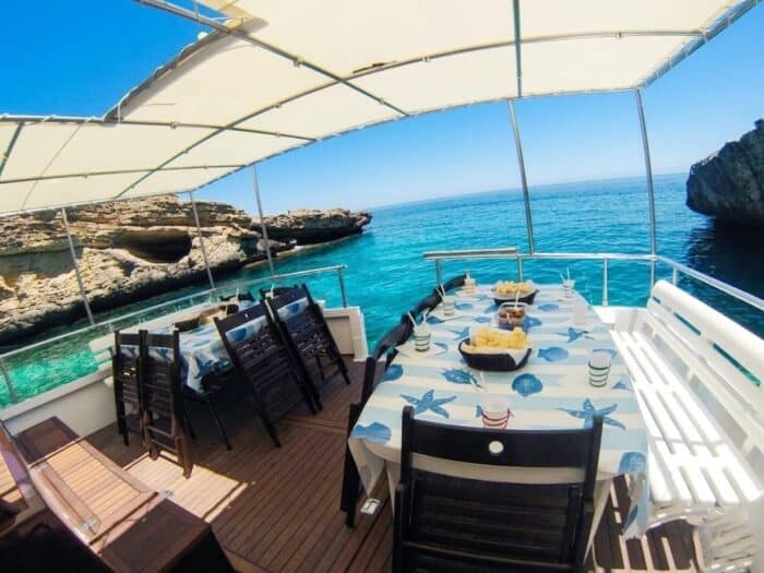 Table set with marine-themed tablecloths and bread baskets on the deck of an excursion boat in Alghero, moored in a cove with turquoise waters and sea caves in the background.