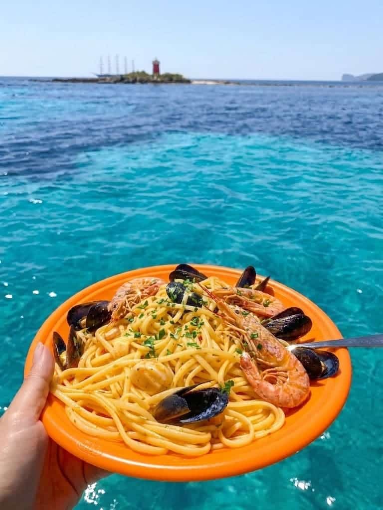 First course of pasta with king prawns and fresh mussels served on board during a boat excursion in Alghero, with the crystalline sea and the lighthouse in the background.