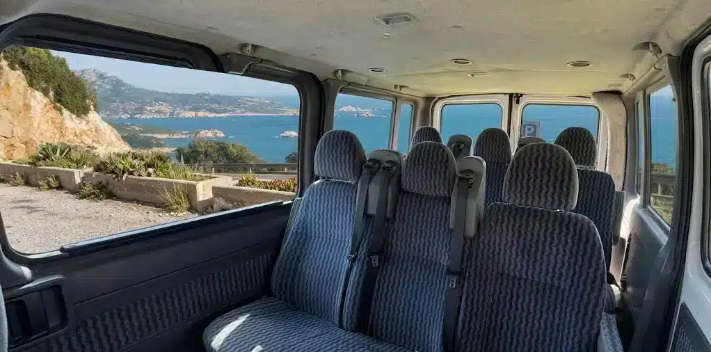 Interior of a passenger van with blue seats and large windows framing a panoramic view of the rocky coast and turquoise sea of Alghero.